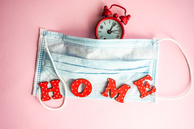 Clock and facemask on pink pastel background with the word "home"