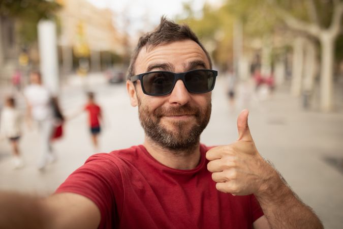A young, bearded man, wearing sunglasses, smiles at the camera, while taking a selfie with his cell phone during a walk in the city