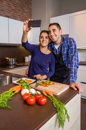 Happy couple in a home kitchen taking a selfie with a smartphone while preparing meal
