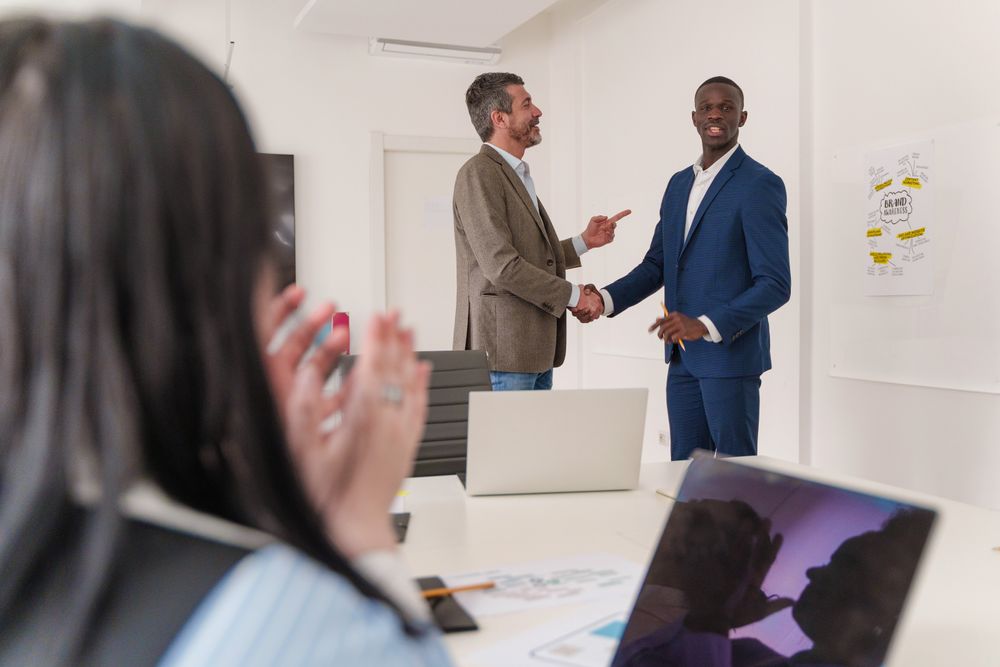 Two businessmen shaking hands in a celebration of a partnership, with colleagues clapping