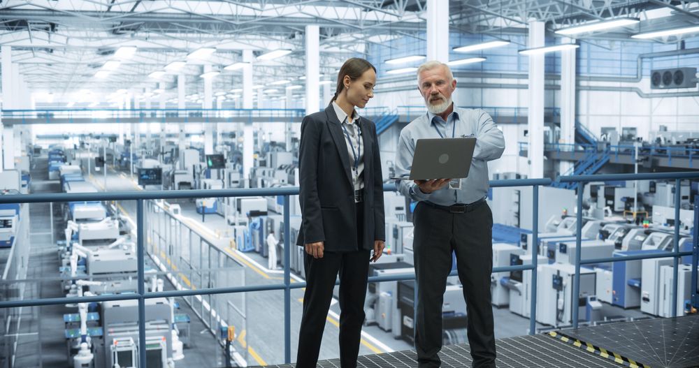 Portrait of a Male and Female Scientist Using Laptop Computer and Talking in a Factory Facility with Equipment Producing Modern Technical Devices for Different Industries