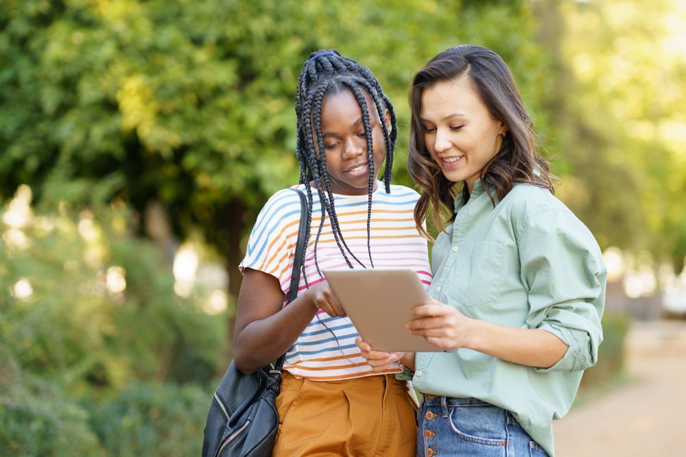 Female friends pointing at something on tablet in casual clothing in park