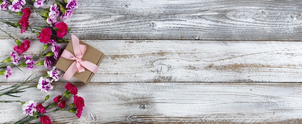 Left side border of carnation flowers and Mothers day gift box on rustic wooden background with copy space. Overhead view in flat lay format