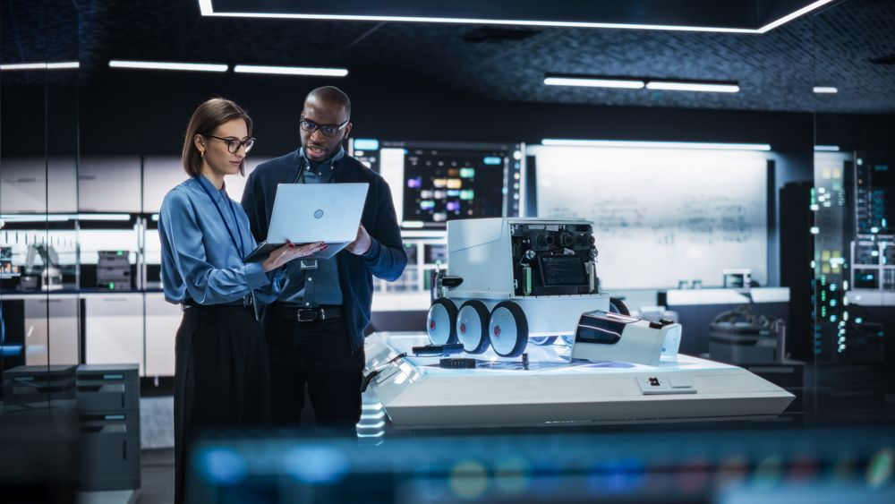 Two Diverse Engineers Developing a Remotely Controlled Mobility Robot in an Industrial Research Facility. Black Male Specialist Pitching his Ideas and Advising his Colleague Using Laptop Computer