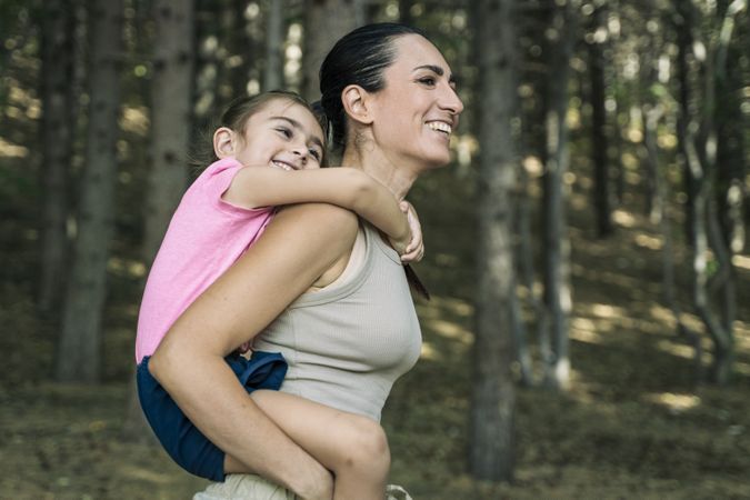 Happy mother giving her smiling daughter a piggyback ride, family having fun in the forest.