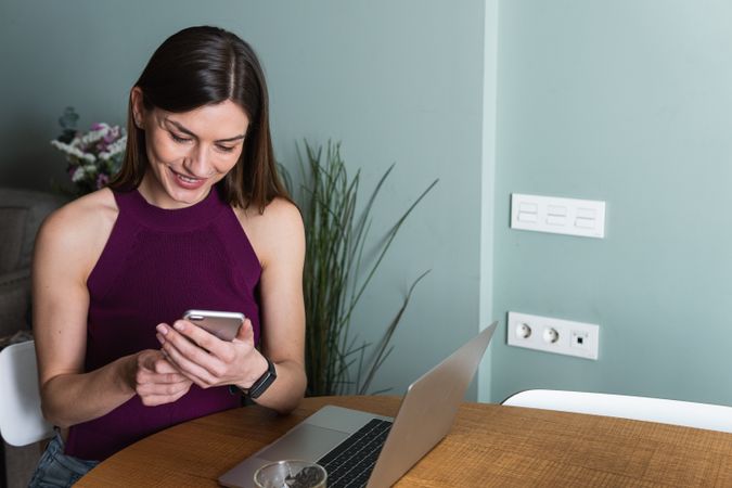 Smiling woman using smartphone and sitting at table with laptop