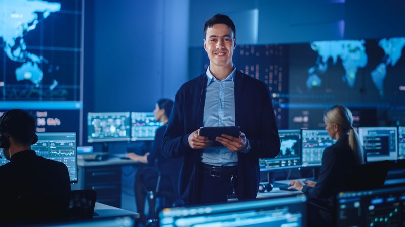 Smiling Professional Holds Tablet in Futuristic Data Center. Team Analyzes Global Information on Screens. Blue-lit Environment With World Map Displays Showcases Advanced Technology.