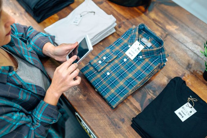 Top view of young female customer taking photo of plaid shirt over counter in clothes shop