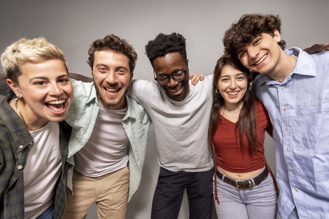 Studio shot of cheerful diverse young adults standing in line and embracing each other with big smiles