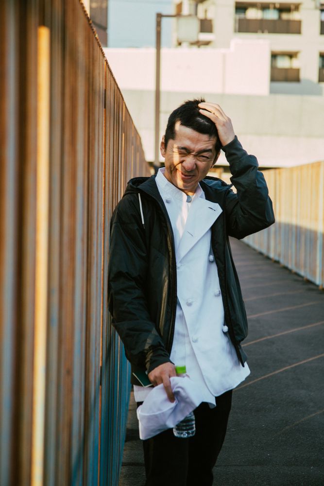 Man in chef uniform and dark jacket standing beside fence