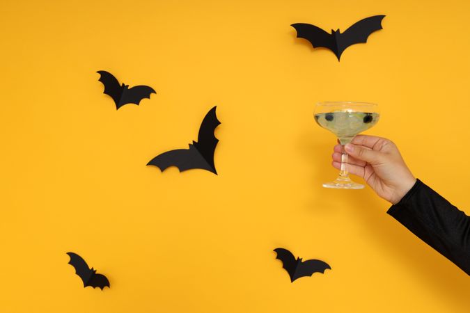 Concept of Halloween, female hand holds cocktail on yellow background with bats