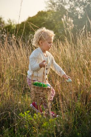 Adorable girl with basket collecting Easter eggs in countryside