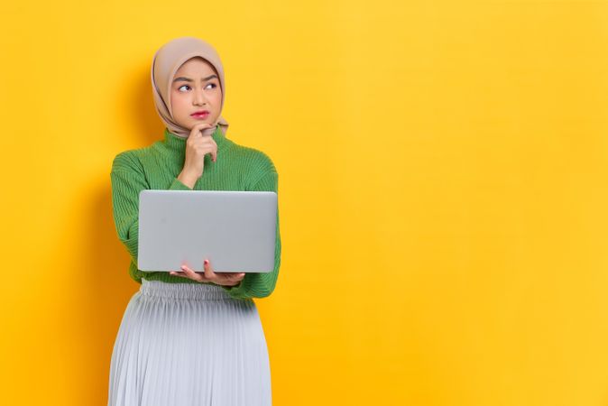Woman in headscarf holding laptop looking up while thinking about something