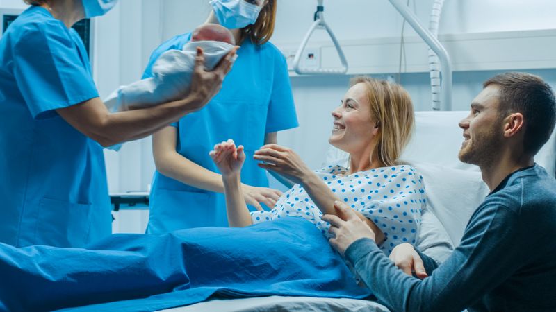 In the Hospital Midwife Gives Newborn Baby to a Mother to Hold, Supportive Father Sitting Near Wife