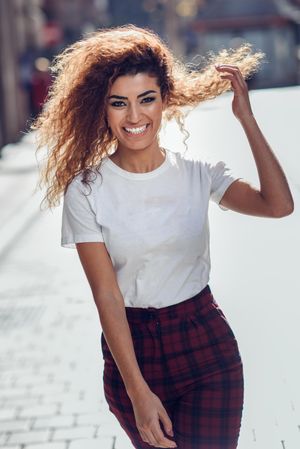 Happy Arab woman with curly hair walking down sunny street playing with hair