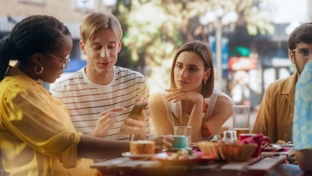 Young Stylish Man Sitting on a Terrace with Multiethnic Friends, Showing Holiday Photos and Social Network Messages on a Smartphone.
