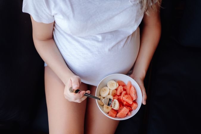 Faceless pregnant woman eating fruits with fork