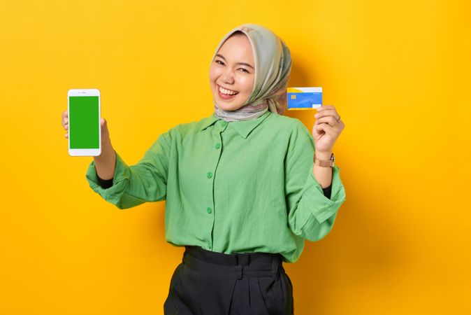 Smiling Muslim woman in headscarf and green blouse with credit card and smart phone screen mock up