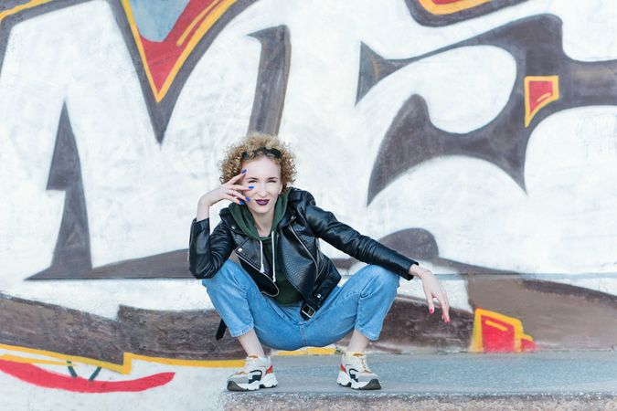 Full length portrait of trendy hipster woman sitting at the graffiti wall background