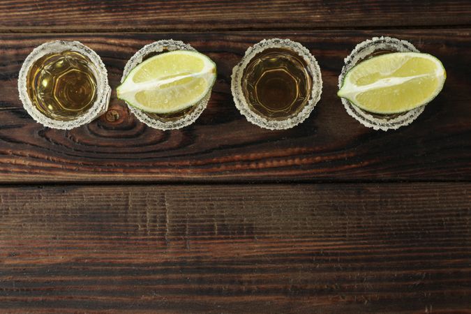 Tequila shots with salt and lime slices on wood table, top view