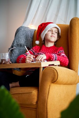 Little dreamy boy in Santa hat painting in armchair