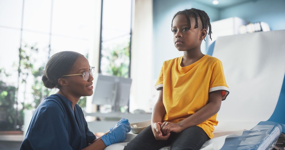 Boy Sitting In The Chair In Hospital, Talking To Female Black Nurse.