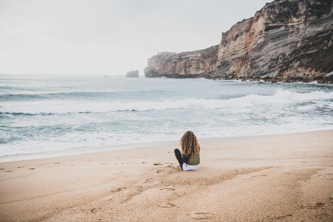 Woman with curly hair sitting on beach in green puffer jacket on fall day