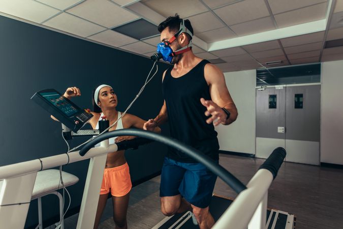 Woman fitness coach monitoring runner with mask on treadmill in laboratory