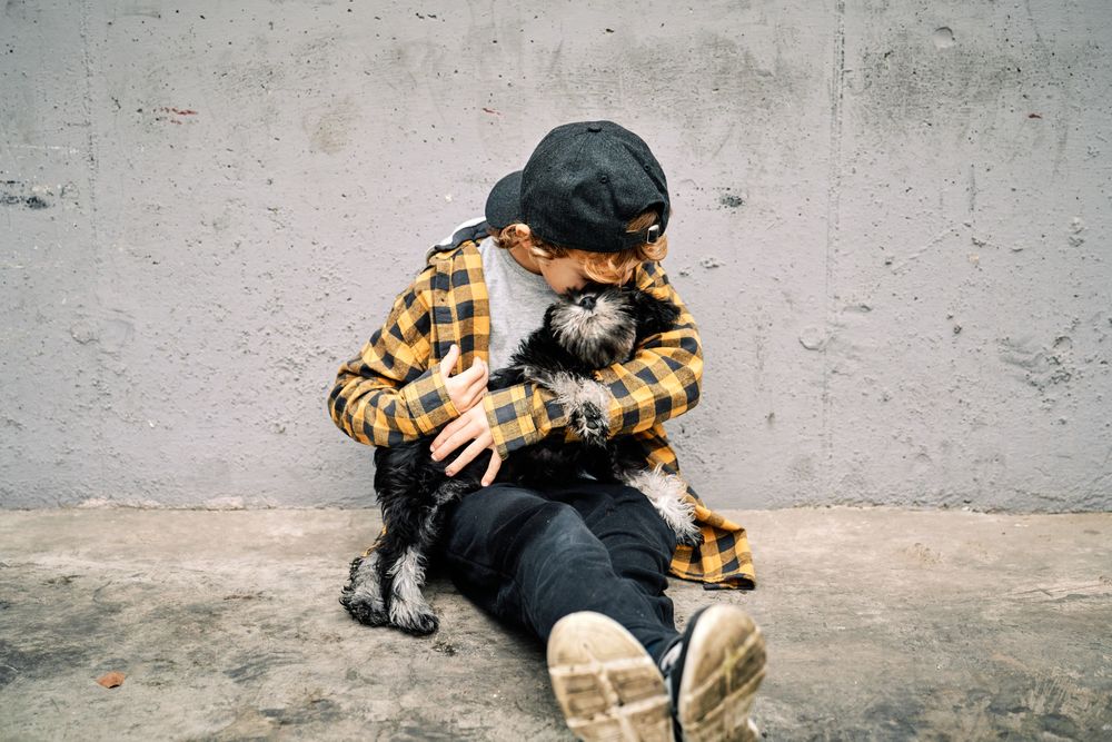 Child hugging dog while sitting near gray street wall