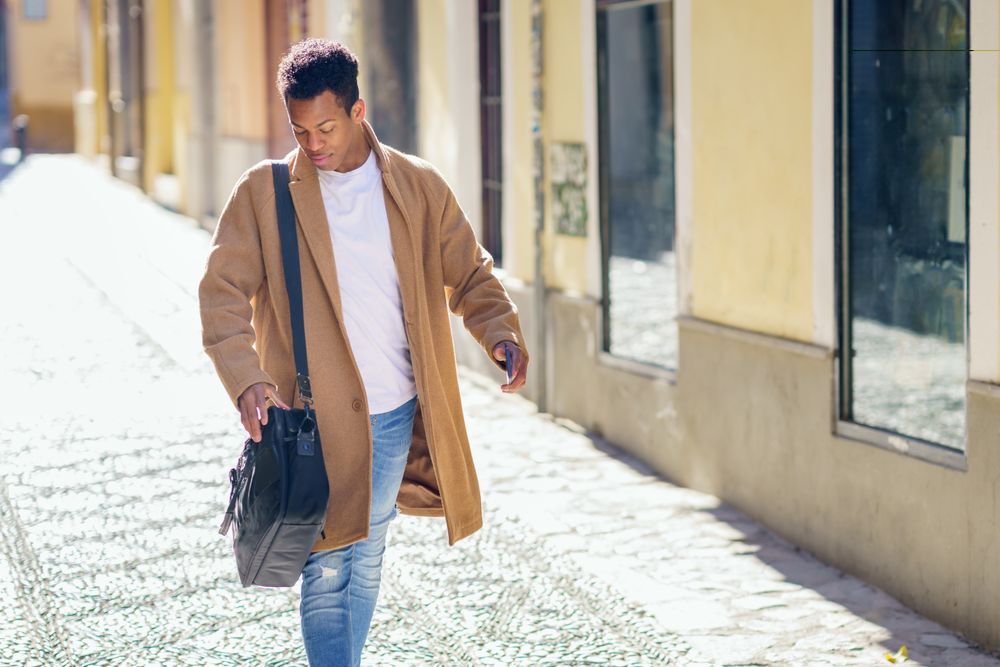 Cuban male strolling down street while looking down, holding leather bag and phone