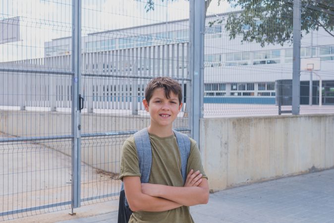 Happy teenage boy standing outside of school entrance with arms crossed