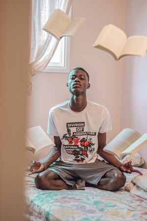 Young man meditating in bedroom with books floating