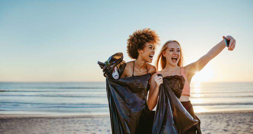Female volunteers with garbage bag taking selfie on the beach after picking up litter