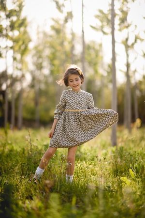 Happy child standing in dress in field surrounded by trees holding dress