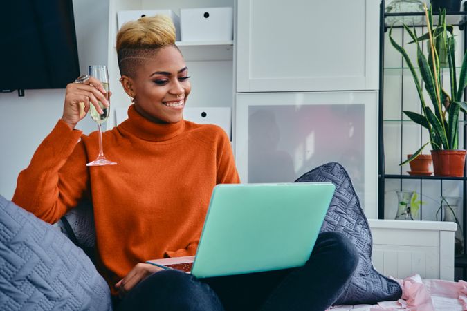 Happy Black woman smiling at a laptop with champagne in a bright modern studio