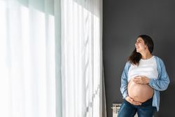 Cheerful pregnant woman standing near window at home