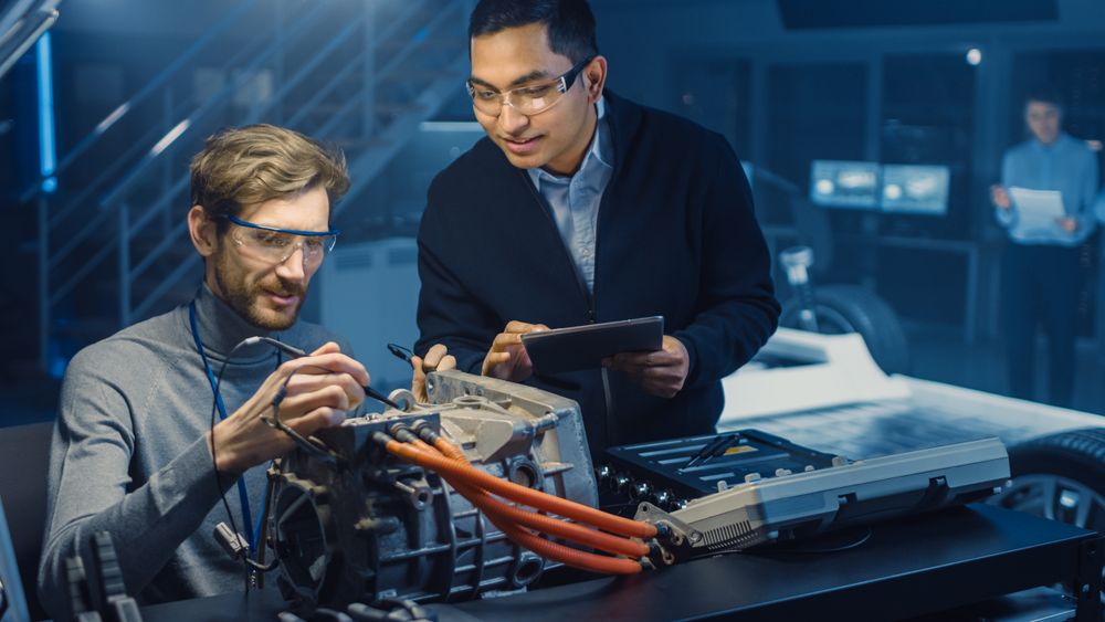 Two Professional Automotive Engineers with a Tablet Computer and Inspection Tools are Having a Conversation While Testing an Electric Engine in a High Tech Laboratory with a Concept Car Chassis.
