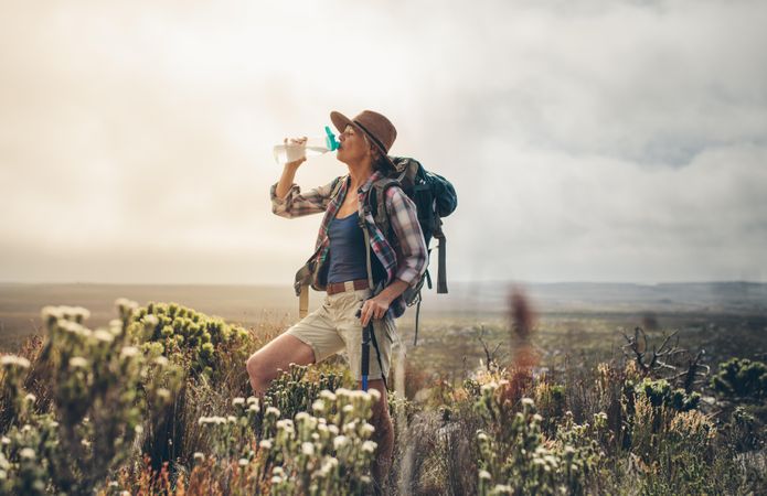 Woman hiker wearing backpack and hat drinking water standing on a hill