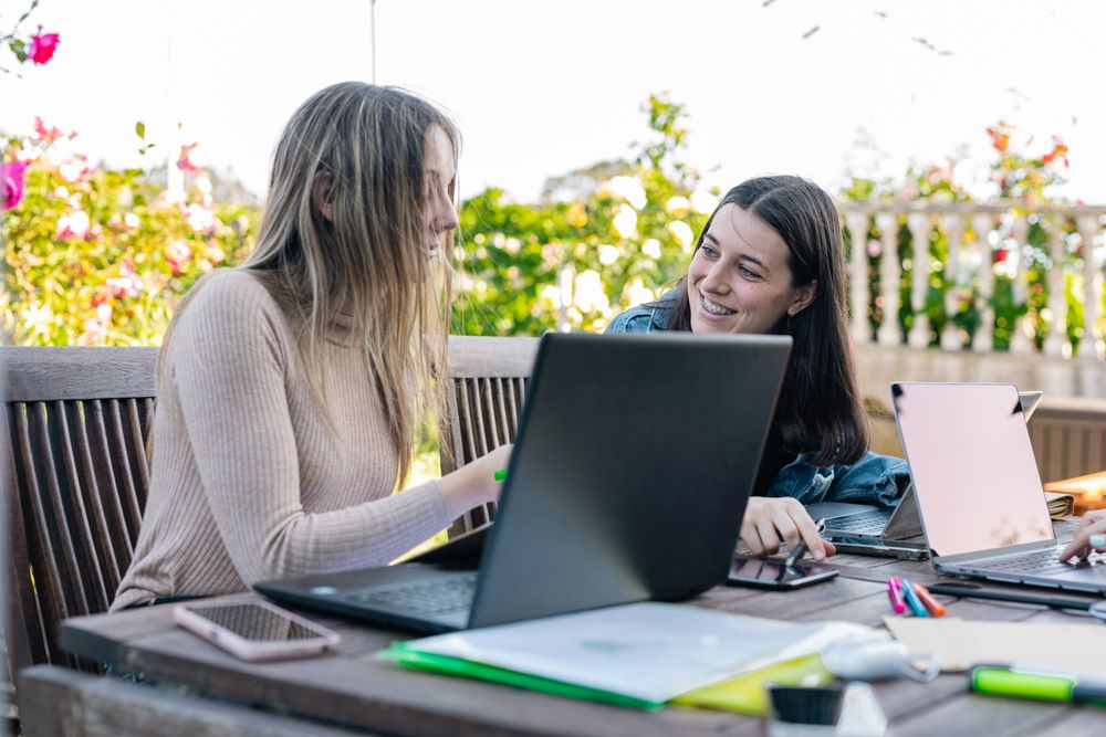 Two smiling female friends comparing their notes and chatting about what was written on the laptop.