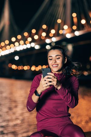 Woman in dark red sportswear smiling at phone near bridge at evening