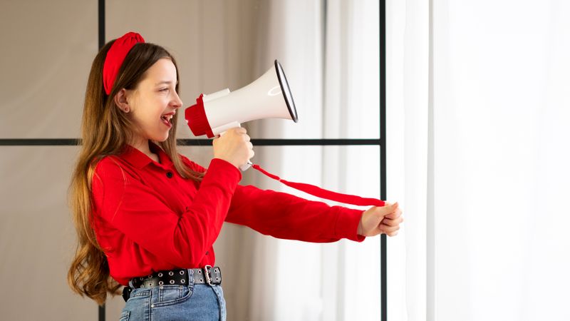 Teen girl speaking into megaphone indoors near window.