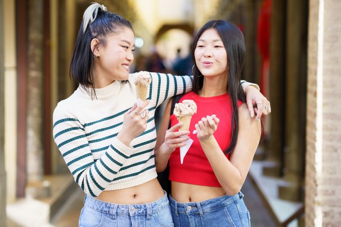 Happy Asian woman standing with hand on shoulder of friend while eating delicious ice cream outside