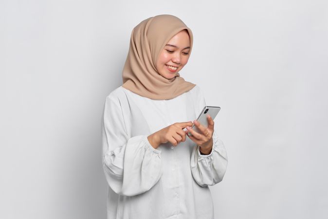 Happy Asian female in headscarf looking down at cell phone and smiling