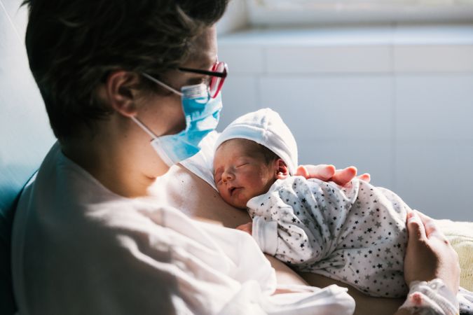 Mother wearing a face mask while holding her newborn baby in the hospital room.
