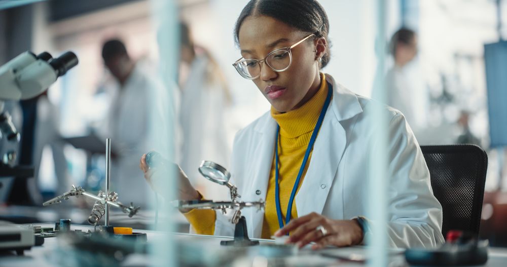 Female Specialist Checking on Electronic Circuit Using Microscope While Team of Professionals