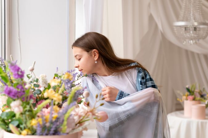 Girl smelling flowers near window with closed eyes indoors.