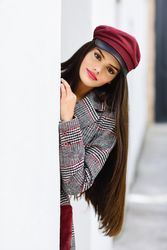 Female in red cap peaking out from behind light wall outdoors, vertical