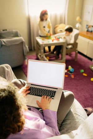 Back view of mother working on her laptop while kids playing in living room