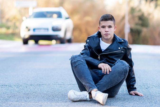 Teenage male wearing a leather jacket sitting crossed legged on concrete with car passing behind him