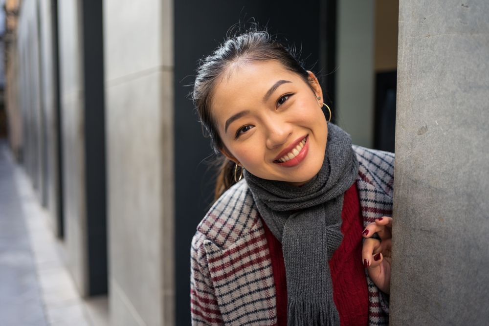 Joyful Asian resting female in casual clothing looking out modern wall of building at city street